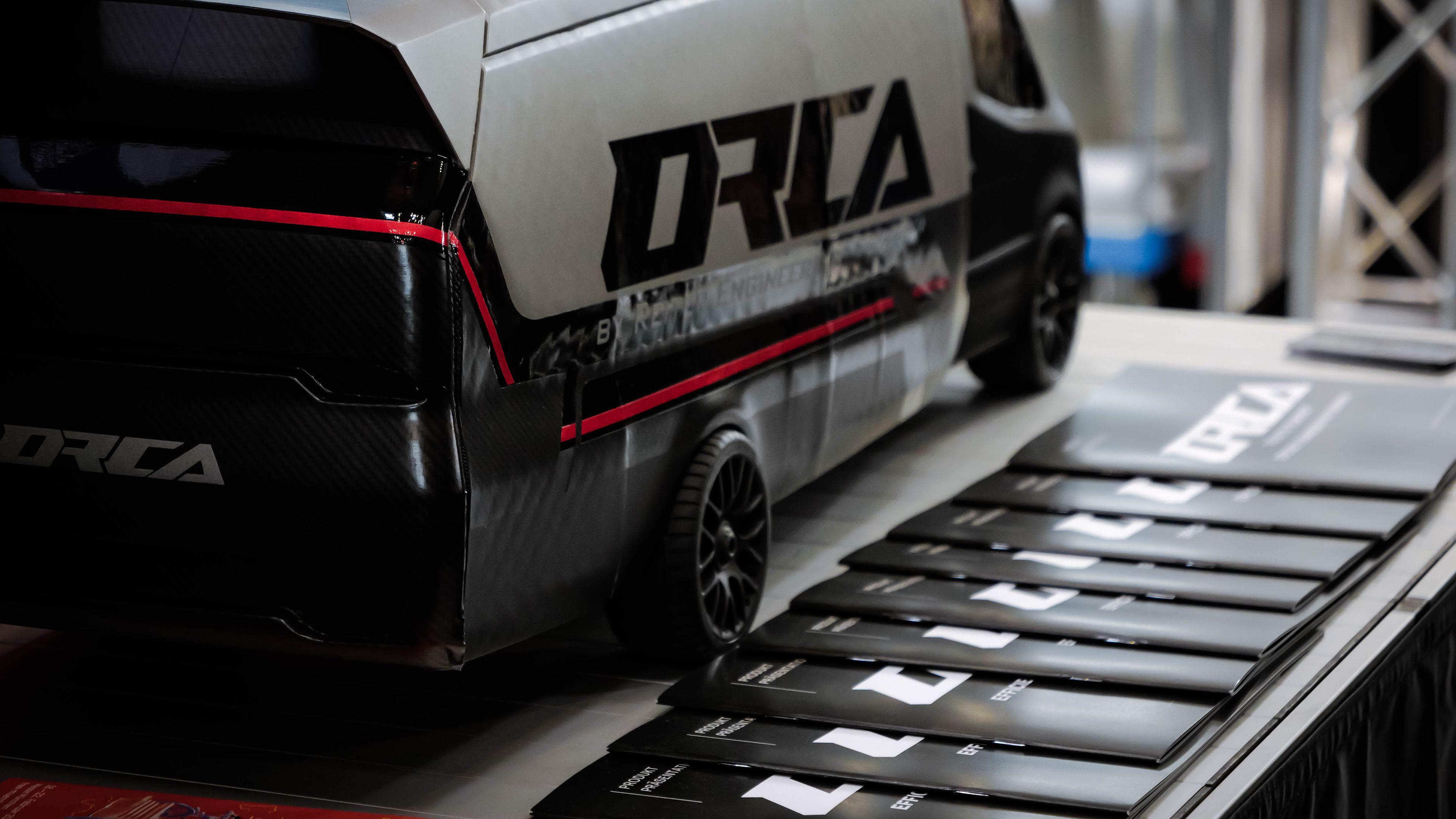 Ein detailliertes Scale-Modell des ORCA Leichtbau-Transporters in Schwarz und Silber mit roten Akzentstreifen, präsentiert auf einem Messestand neben Informationsbroschüren