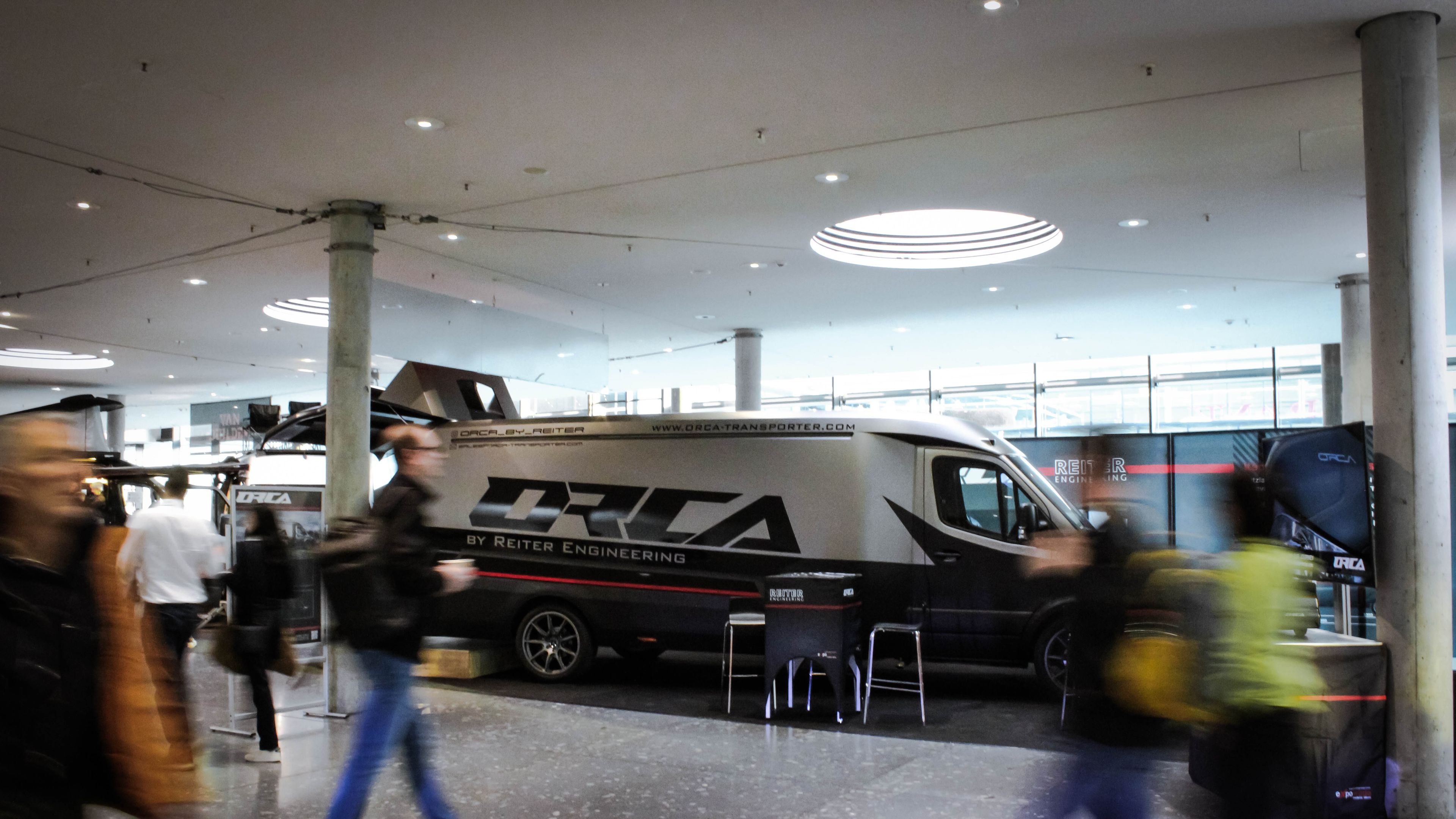 Seitenansicht des silber-schwarzen ORCA Transporters von Reiter Engineering auf einer Messe mit Besuchern.