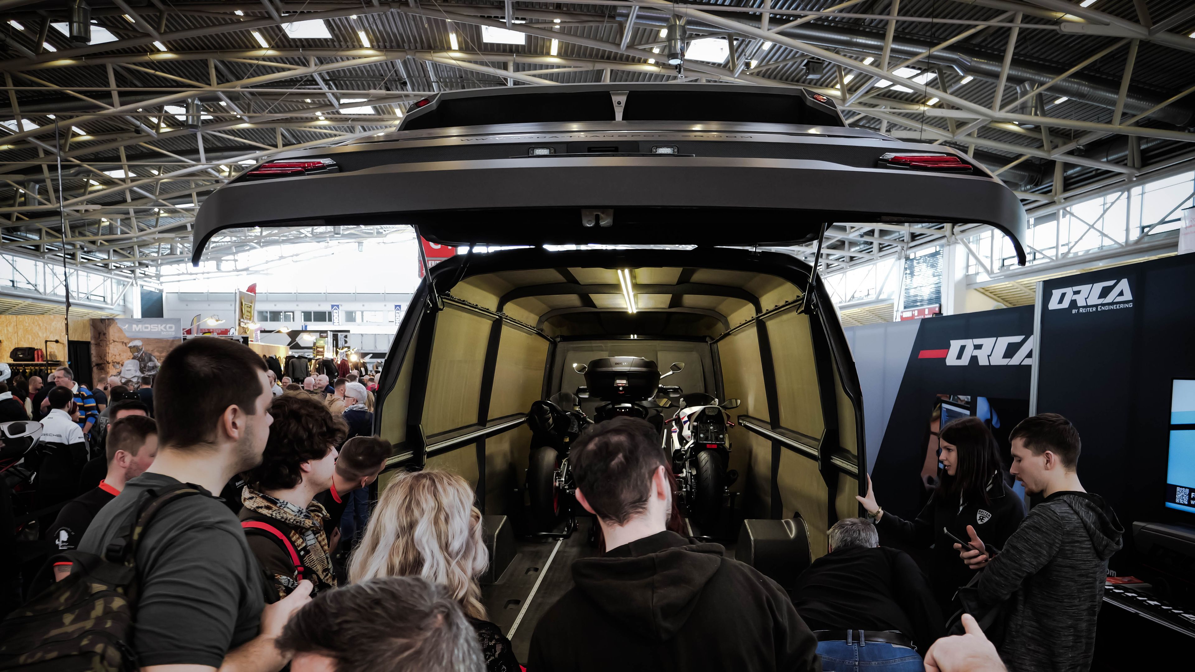 Eine Menschenmenge begutachtet das Heck des ORCA Leichtbau-Transporters bei einer Messe. Die Heckklappe ist geöffnet und gibt den Blick auf mehrere verladene Motorräder im Innenraum frei.